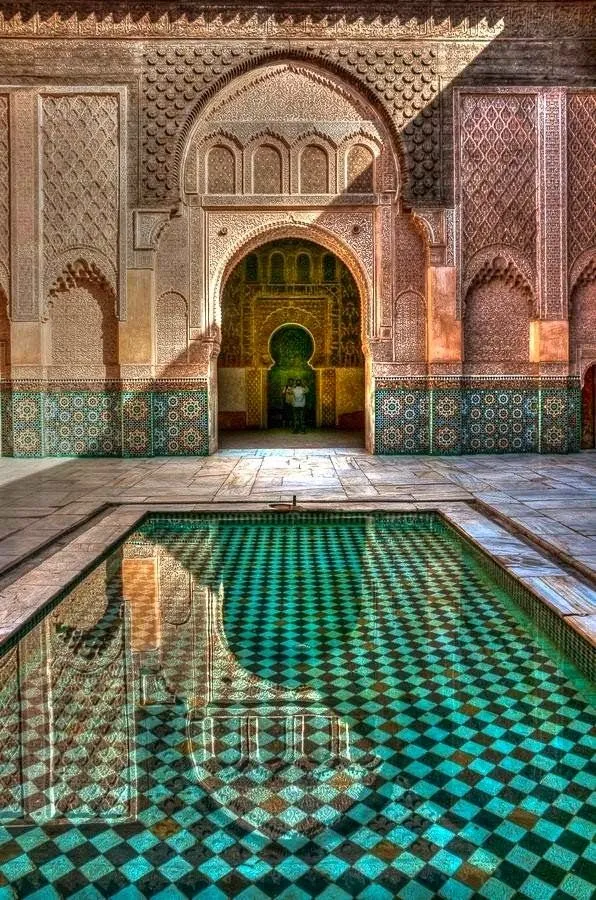 The courtyard of Medersa Ben Youssef in Marrakesh, with a reflective pool of turquoise and black checkered tiles, surrounded by intricate stucco work, carved arches, and walls with geometric patterns and zellige tilework, showcasing Moroccan architecture.