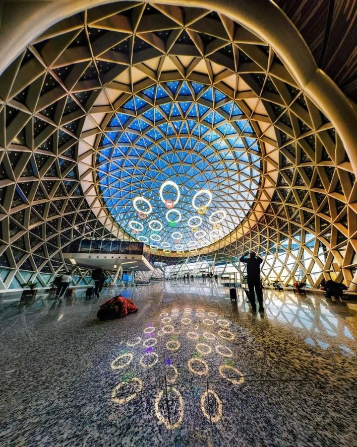 The modern interior of Menara Airport in Marrakesh, with a lattice ceiling showcasing a blue, starry design and circular lights. The polished floor reflects the patterns, and travelers are visible in the spacious terminal.