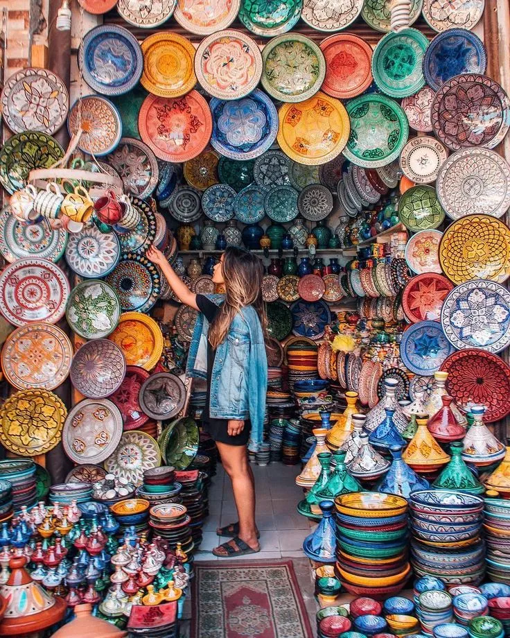 A person browses a vibrant pottery shop in a Marrakesh souk, surrounded by colorful plates, bowls, and tagines in shades of blue, red, yellow, and green, displayed on the walls and floor of the traditional market.