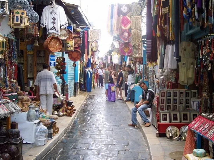 A lively alley in a Casablanca souk, a must-visit in Casablanca, with cobblestone paths lined with stalls selling colorful textiles, lanterns, pottery, and souvenirs, as locals and tourists explore the vibrant market, a top Casablanca attraction.