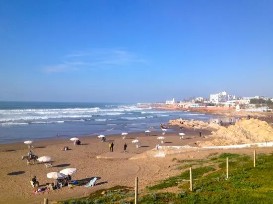 Ain Diab Beach in Casablanca, a coastal gem to visit in Casablanca, featuring a sandy shoreline with waves, umbrellas, and beachgoers under a clear blue sky, with the city skyline in the background, a top Casablanca attraction.