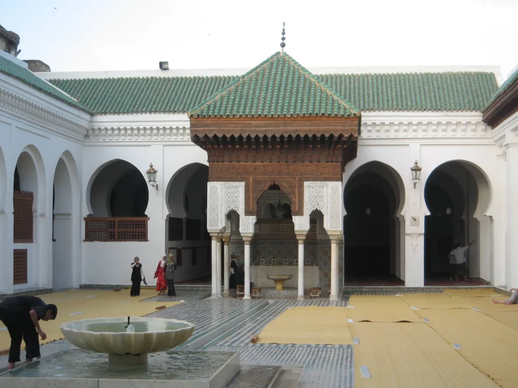 The courtyard of Al Quaraouiyine Mosque and University, a historic site for things to do in Fes, with a marble fountain, arched doorways, intricate tilework, a green-tiled roof, and visitors on woven mats, a top Fes Morocco attraction.