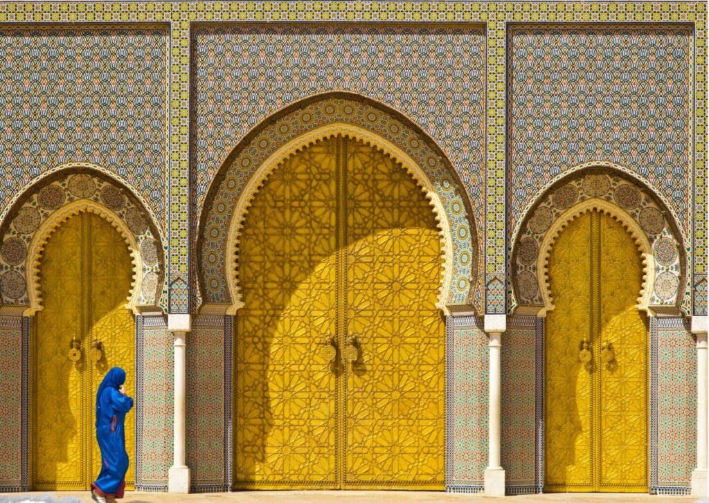 The ornate Gates of the Royal Palace in Fes, a stunning attraction for things to do in Fes, featuring golden doors with geometric patterns and colorful zellige tilework, with a person in a blue robe nearby, a top Fes Morocco attraction.