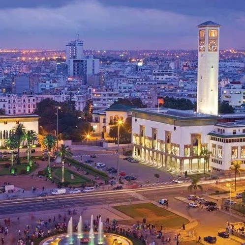 Mohammed V Square in Casablanca at dusk, a top spot to visit in Casablanca, with a lit fountain, palm trees, and crowds, featuring the Wilaya building and clock tower against the city skyline, a key Casablanca attraction.
